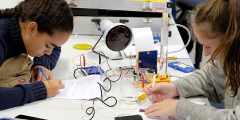 Das Foto zeigt zwei Mädchen an einem Tisch, die Berechnungen auf einem Blatt Papier durchführen. Auf dem Tisch stehen verschiedene Teile eines Experiments, wie Glasgefäße und Kabel.