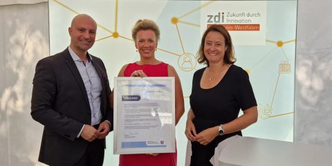 Das Foto zeigt Landrat Ali Doğan, Ministerin Ina Brandes und Regierungspräsidentin Anna Katharina Bölling. Die Ministerin hält eine Urkunde zur Ernennung des zdi-Schüler:innenlabors in der Hand. Die drei stehen vor einer Leuchtwand, auf der zdi - Zukunft durch Innovation Nordrhein-Westfalen steht.