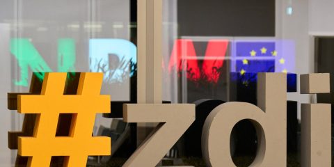 Ein Schild mit der Aufschrift „zdi“ vor einem Fenster. Durch das Fenster sieht man die Buchstaben NRW in Grün, Weiß und Rot und eine Europafahne leuchten.