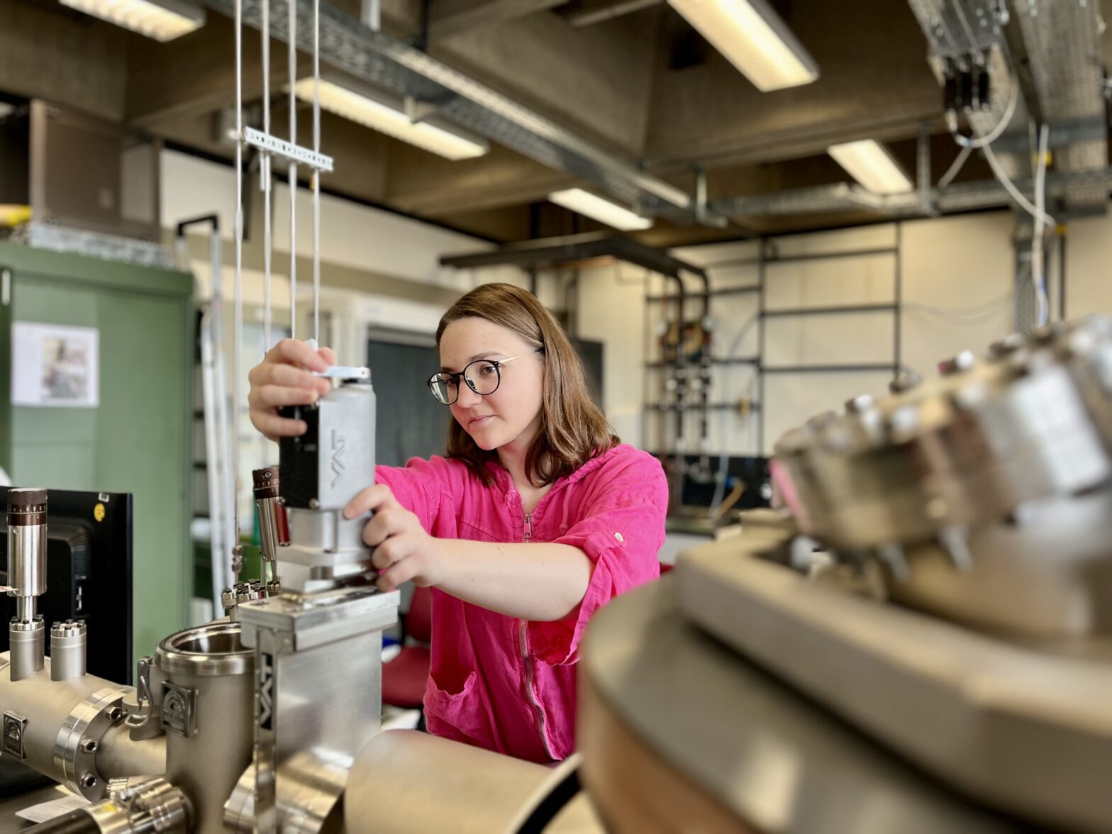 Eine junge Frau mit halblangem Haar, Brille und pinkem Shirt steht an einer naturwissenschaftlichen Apparatur, an der sie arbeitet.