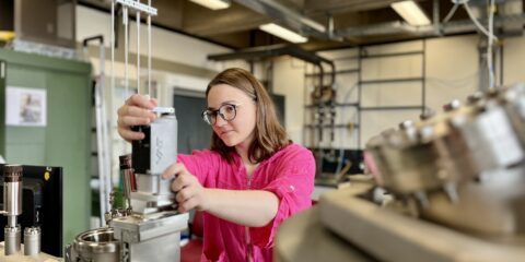 Eine junge Frau mit halblangem Haar, Brille und pinkem Shirt steht an einer naturwissenschaftlichen Apparatur, an der sie arbeitet.