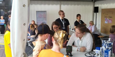 Menschen unterhalten sich an einem Stand in einem weißen Zelt bei einer Veranstaltung. Eine Frau und ein Kind, von hinten zu sehen, unterhalten sich mit Mitarbeitern hinter einem Tisch mit der Aufschrift „Westfalen“, auf dem verschiedene Gegenstände und ein Schild ausgestellt sind. Die lebhafte Szene ist Teil der Feierlichkeiten zum NRW-Tag 2024.