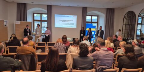 Eine Gruppe von Menschen in einem Konferenzraum hört sich einen Vortrag zum Thema „Zukunftsberufe für Mädchen“ an. Vorne stehen mehrere Vortragende, hinter ihnen läuft eine projizierte Präsentation. Diese Veranstaltung des zdi-Heldinnen-Oktobers fesselt die Anwesenden, die den Vortragenden gegenübersitzen.