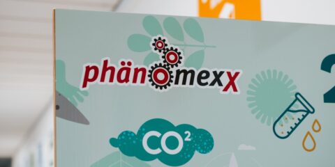 Das Bild zeigt ein Schild mit dem Text „PhänomexX Krefeld“, begleitet von Abbildungen von Zahnrädern, einem CO₂-Symbol, einem Reagenzglas und einem Tropfen auf hellem Hintergrund.