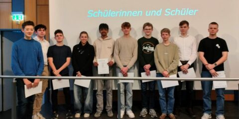 Zehn Schüler:innen stehen vor einem Bildschirm auf dem steht "Die Gruppen der Schülerinnen und Schüler". Sie halten jeweils ein Blatt Papier in der Hand und posieren versetzt in einer Reihe.