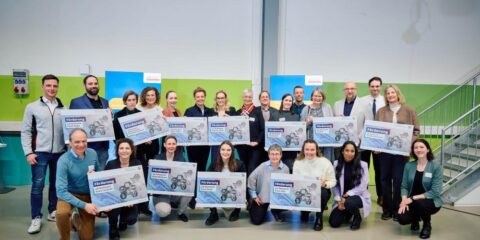 Eine Gruppe von Menschen steht in einem Gebäude, hält in zwei Reihen große Schilder in den Händen und lächelt in die Kamera.